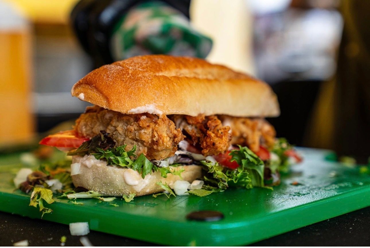 A fried chicken sub with greens and tomato on a green board