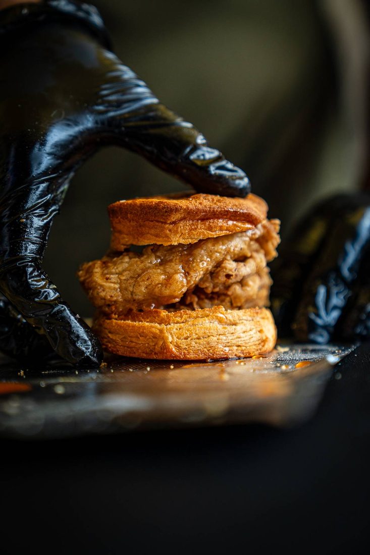 A fried chicken biscuit sandwich pressed by a gloved hand